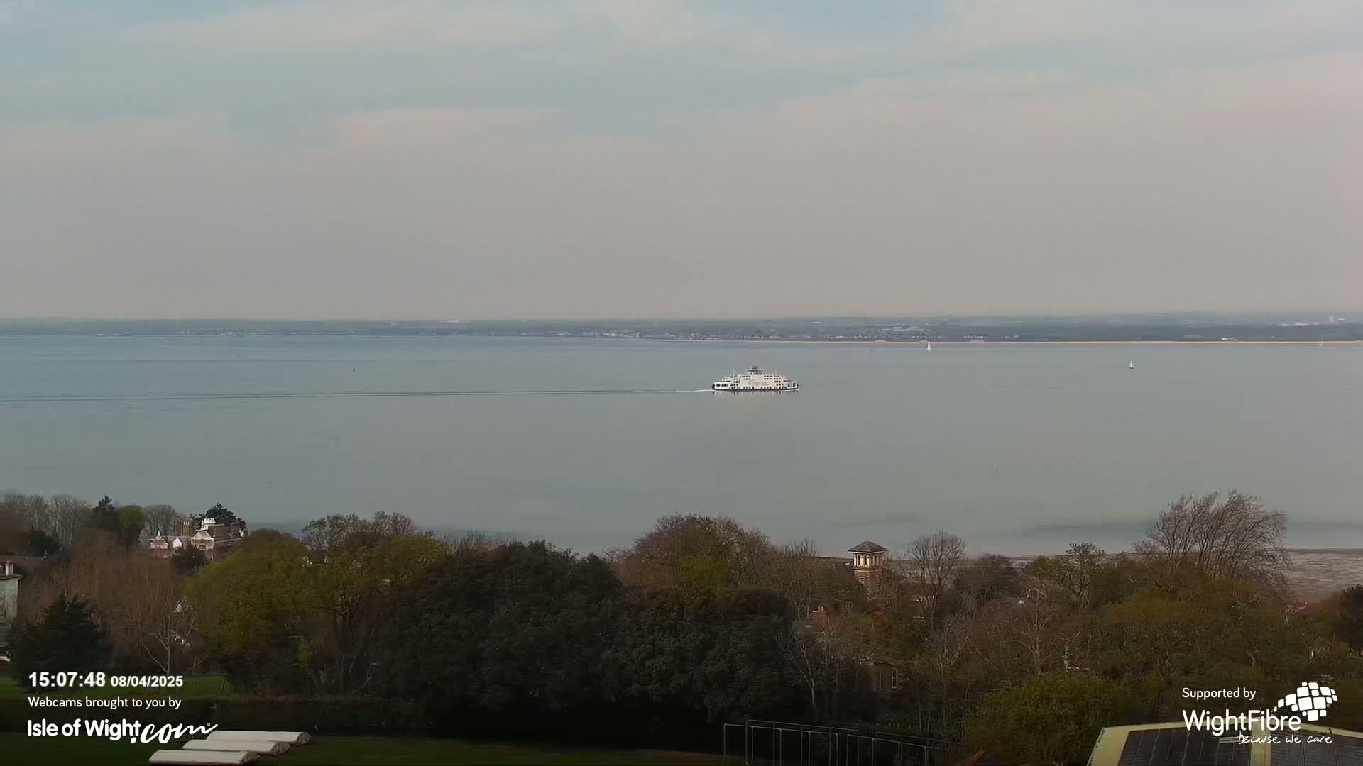 A ferry travels across a calm, gray body of water under a cloudy sky, with trees and buildings visible in the foreground.