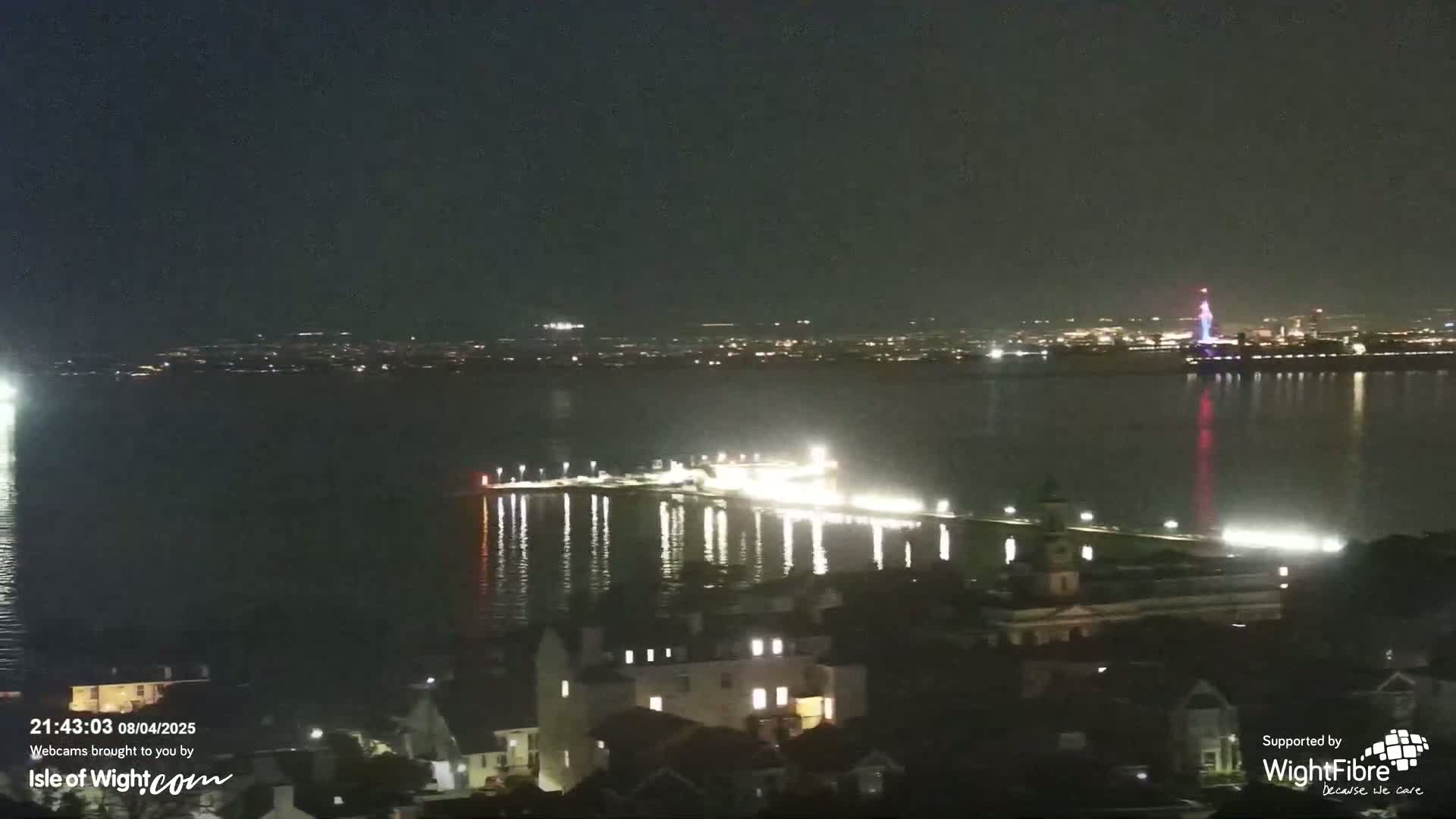 A brightly lit pier extends into a calm, dark body of water at night, with a city skyline visible in the distance.