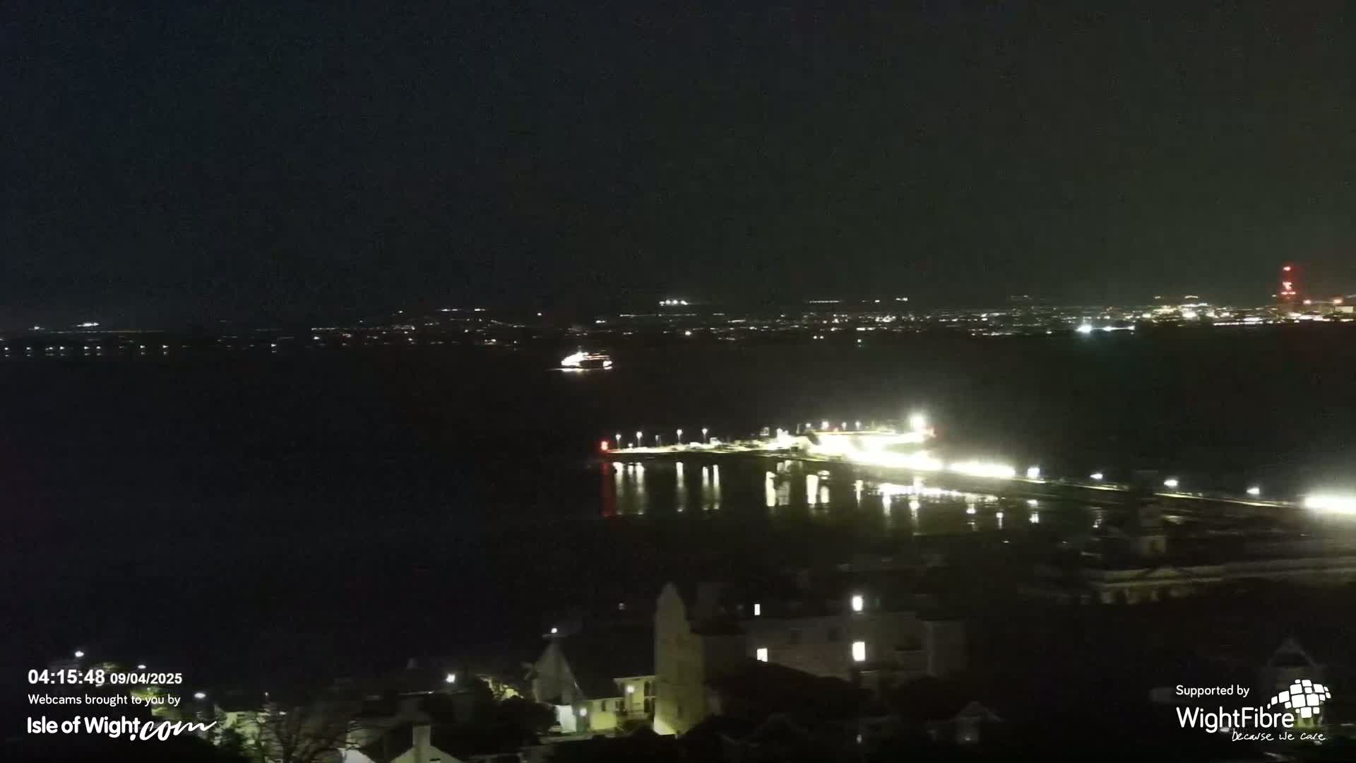 A nighttime view of a body of water with a lit-up pier and distant city lights under a dark sky.