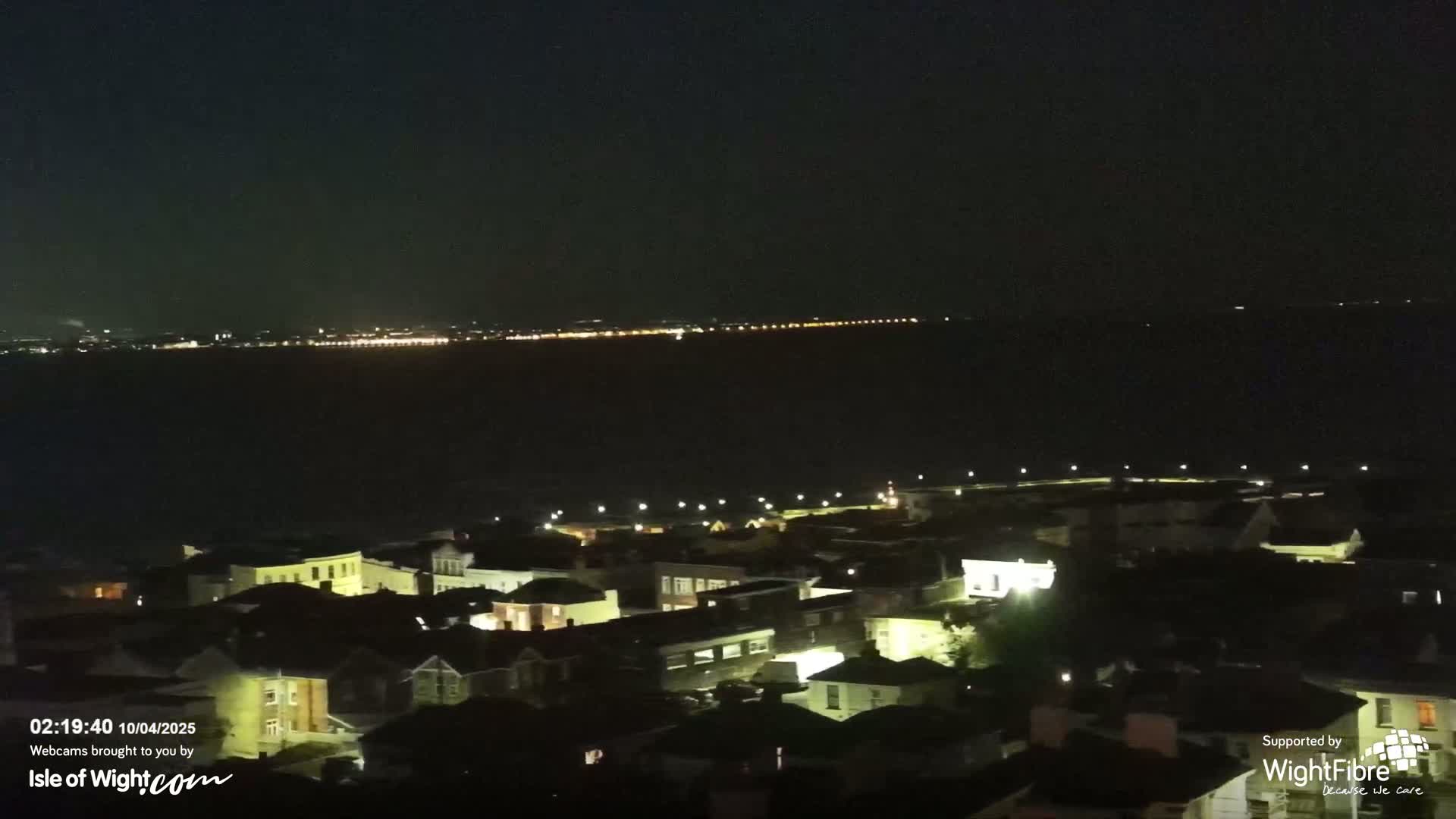 A nighttime view shows a town's illuminated rooftops and streets overlooking a dark body of water with a distant, faintly lit coastline.
