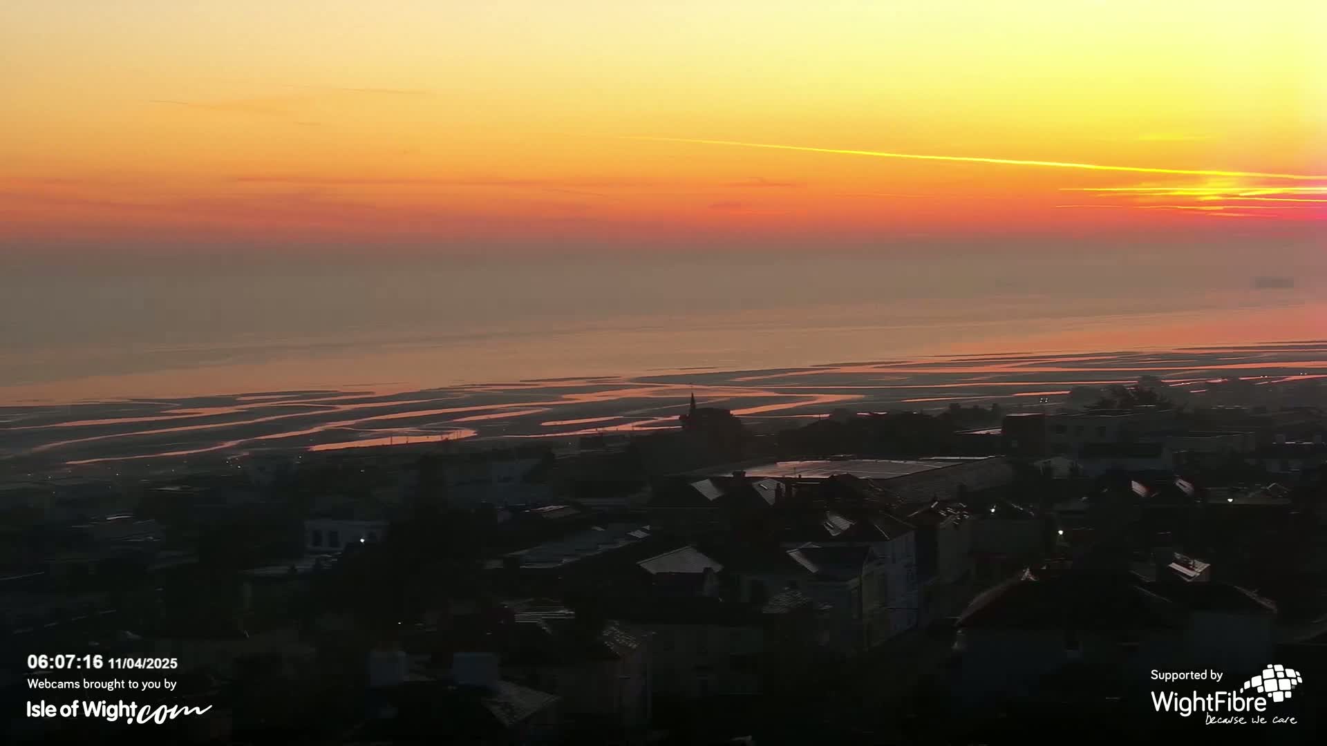 A coastal town at sunset is viewed from above, showing a calm sea with tidal flats and an orange and pink sky.