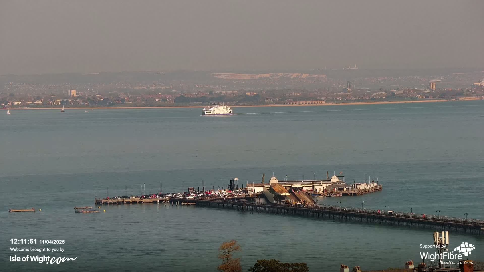 A ferry crosses a calm, hazy body of water toward a long pier with cars parked on it.