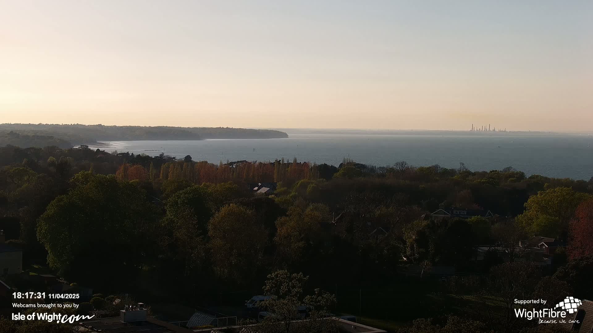 A calm body of water stretches to a distant industrial skyline under a pale, sunset sky, viewed from an elevated vantage point overlooking a tree-lined shoreline and residential area.