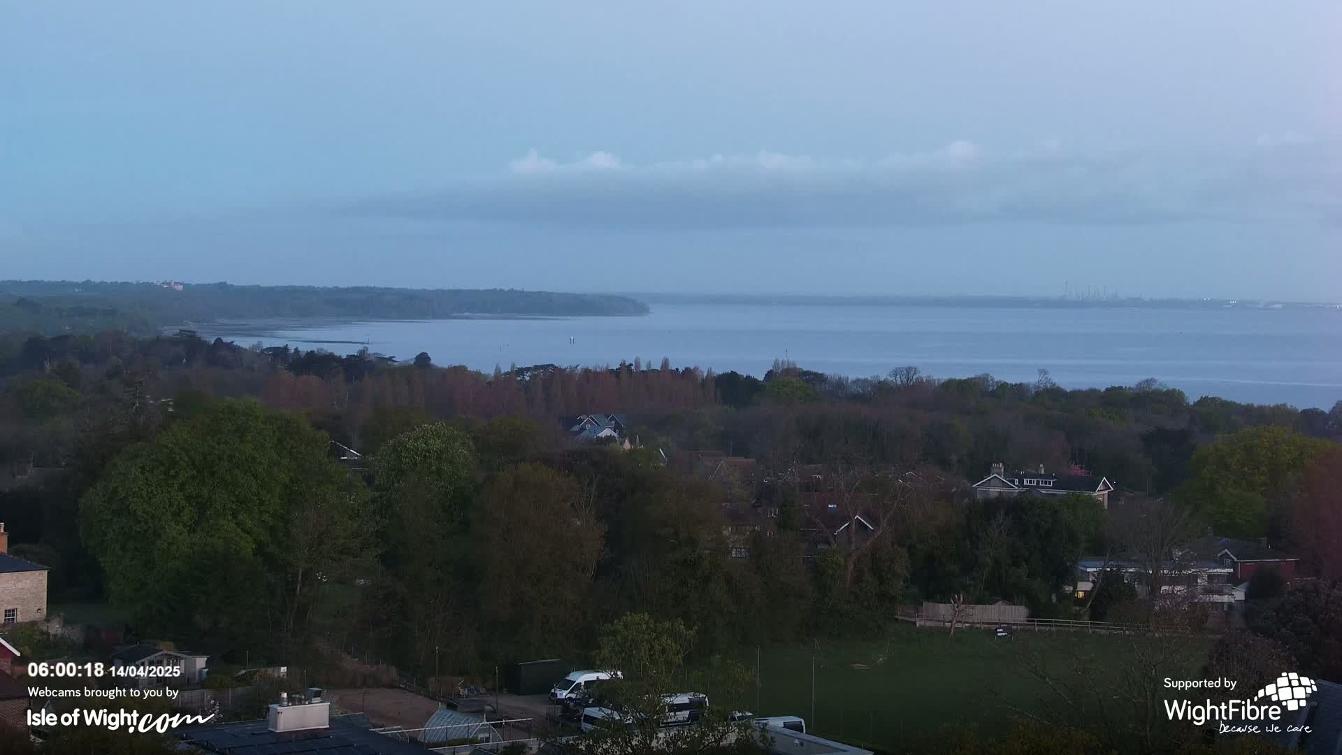 A high-angle view of a tranquil residential area nestled among lush greenery overlooks a calm body of water under a slightly cloudy, dusky sky.