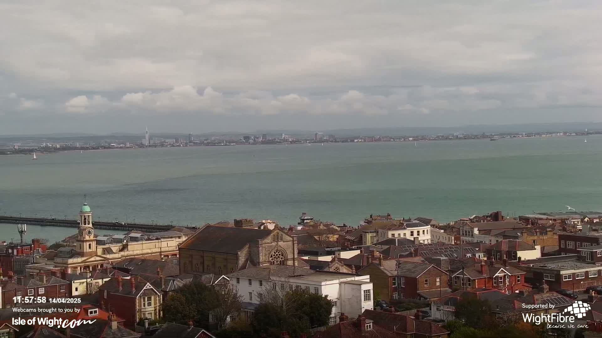A coastal town with a church and many buildings is seen under a cloudy sky, overlooking a body of water and a distant city skyline.