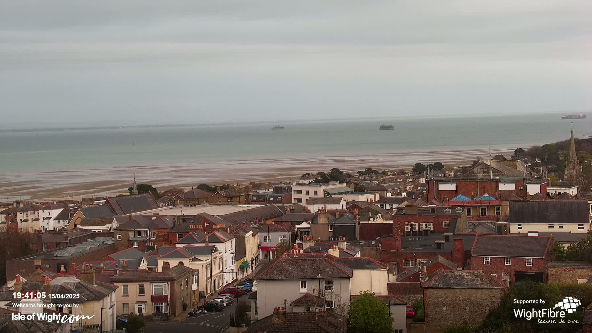 An overcast day reveals a coastal town with tightly packed houses and buildings, overlooking a wide expanse of calm water and exposed sand at low tide, with a few distant ships visible.