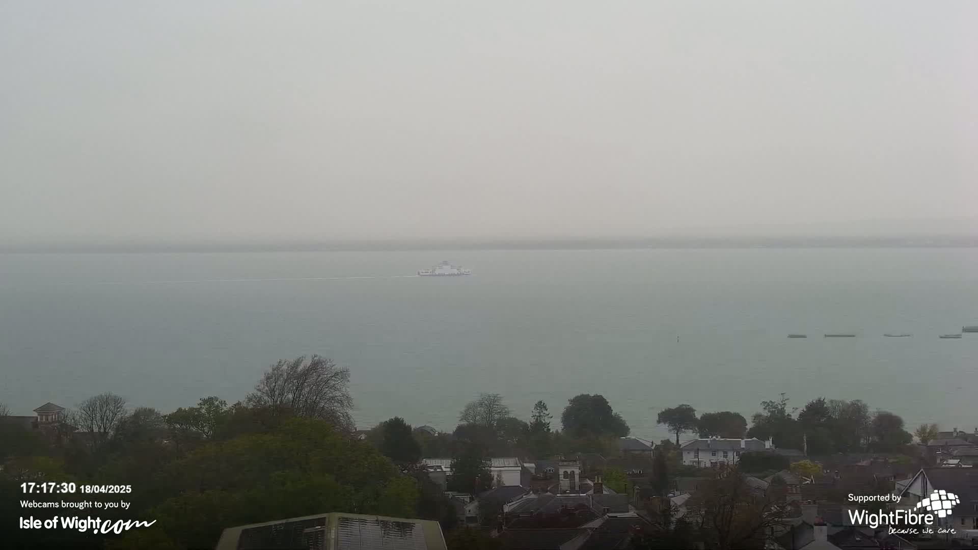 A ferry travels across a calm, hazy body of water, with a residential area and trees visible in the foreground.