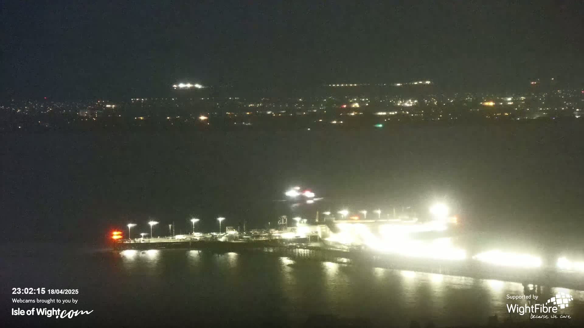 A brightly lit pier extends into dark water at night, with a distant city skyline visible under a cloudy sky.