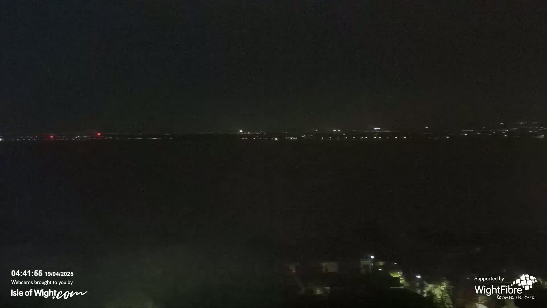A nighttime view of a dark body of water with faintly visible lights on the opposite shore under a clear, dark sky.