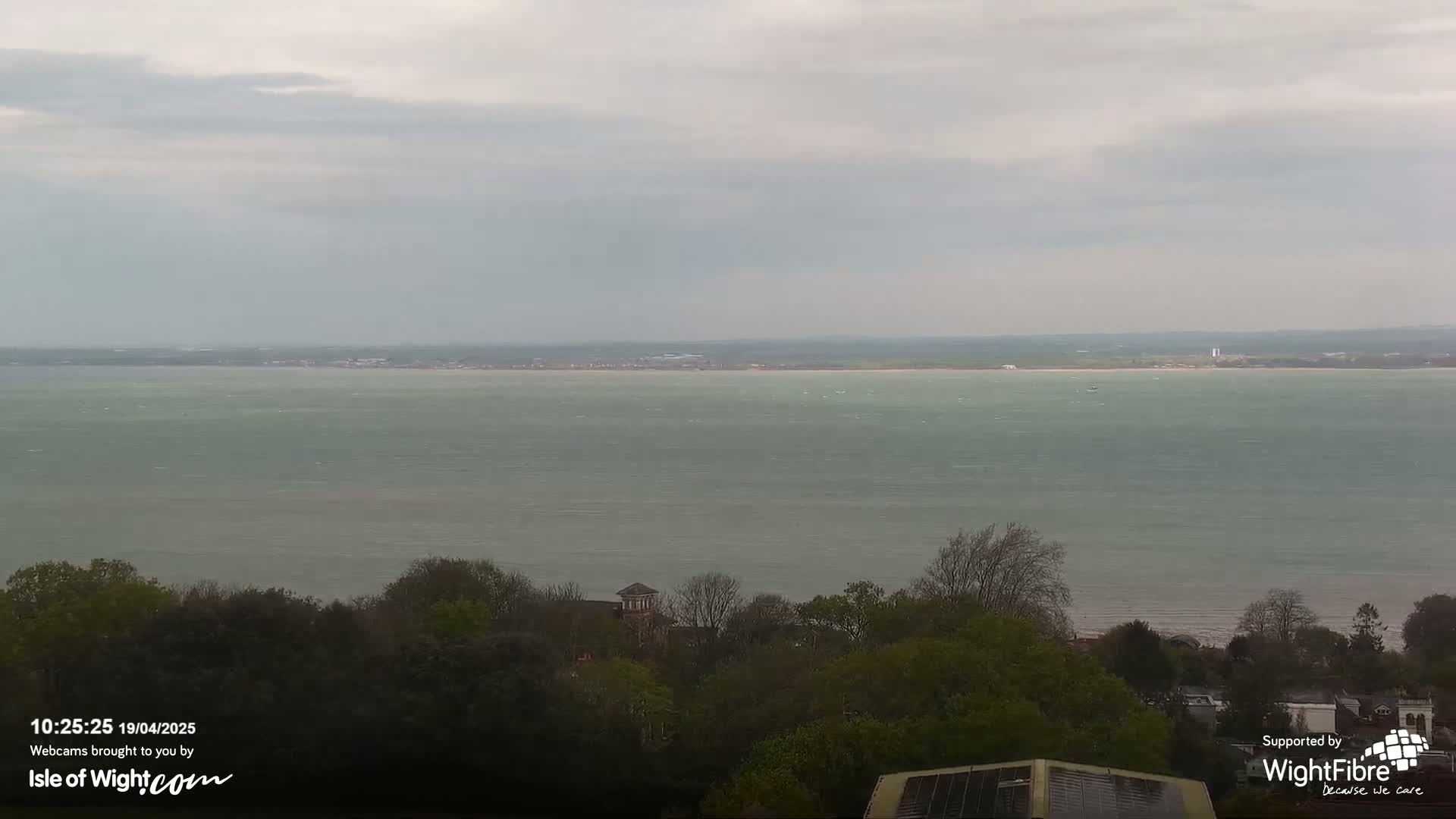 A large body of water stretches to the horizon under an overcast sky, with green trees and buildings visible in the foreground.