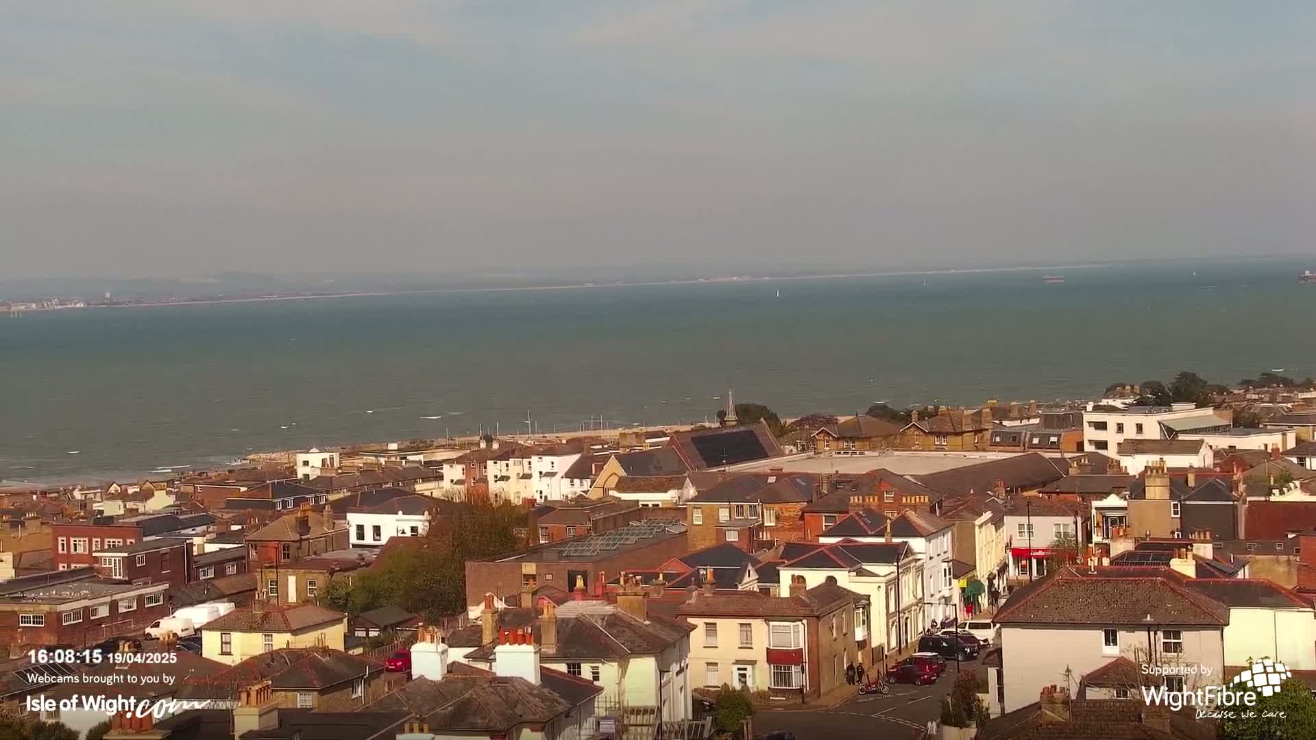 A coastal town with mostly brick houses sits below a calm body of water under a mostly clear sky.