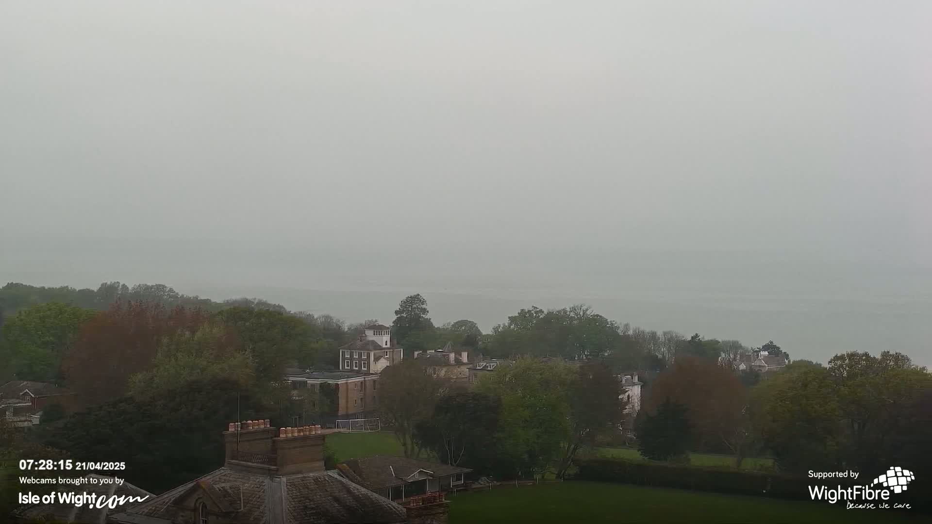 A hazy view of a body of water and tree-lined landscape with several buildings, including a large house, visible in the mid-ground.