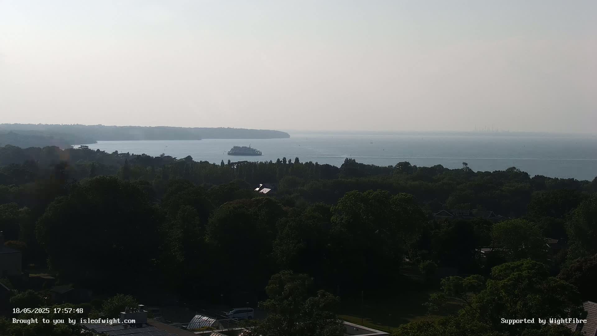 A ferry is visible on a calm, hazy body of water beyond a dense, dark green forest.