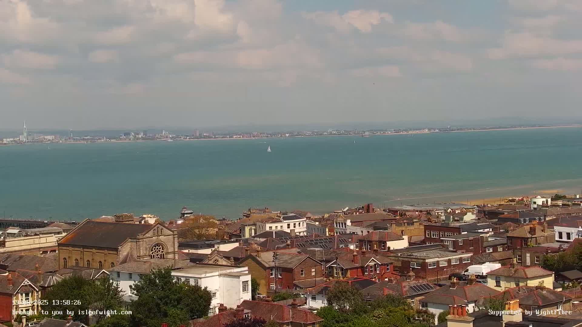 A coastal town with red-tiled rooftops overlooks a calm, teal sea under a partly cloudy sky.