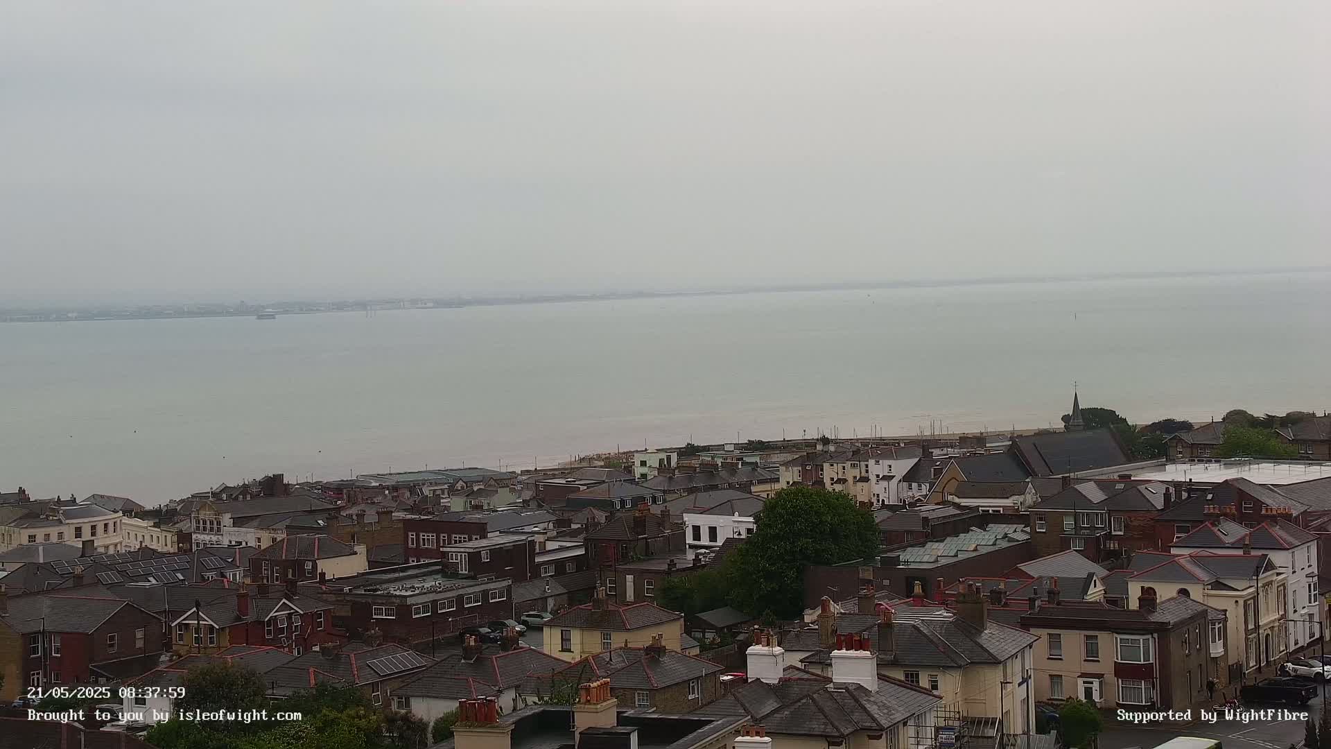 A coastal town with many houses and a calm, grey sea stretches out in the distance under an overcast sky.