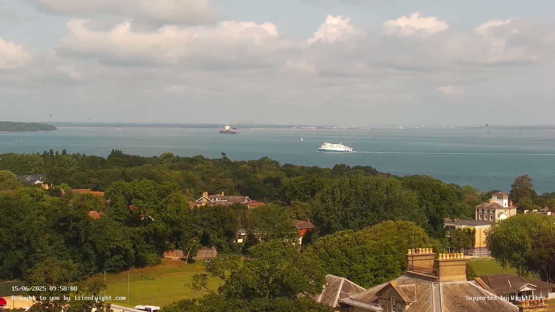A large body of water with a ferry and cargo ship is seen in the distance, beyond a tree-lined hilltop with several buildings, under a partly cloudy sky.