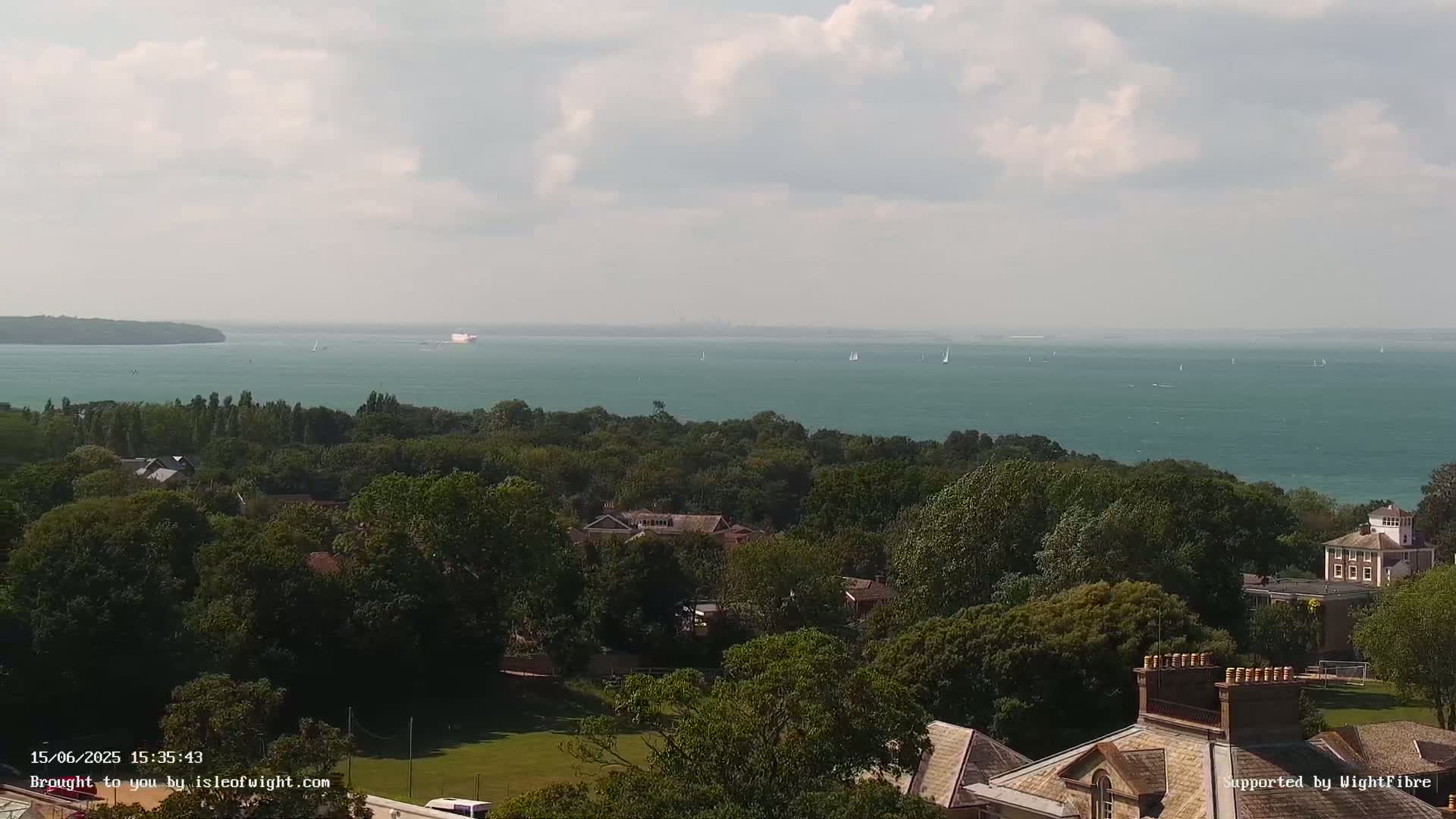 A panoramic view shows a calm body of water with several sailboats, an island in the distance, and a lush green landscape dotted with buildings in the foreground under a partly cloudy sky.