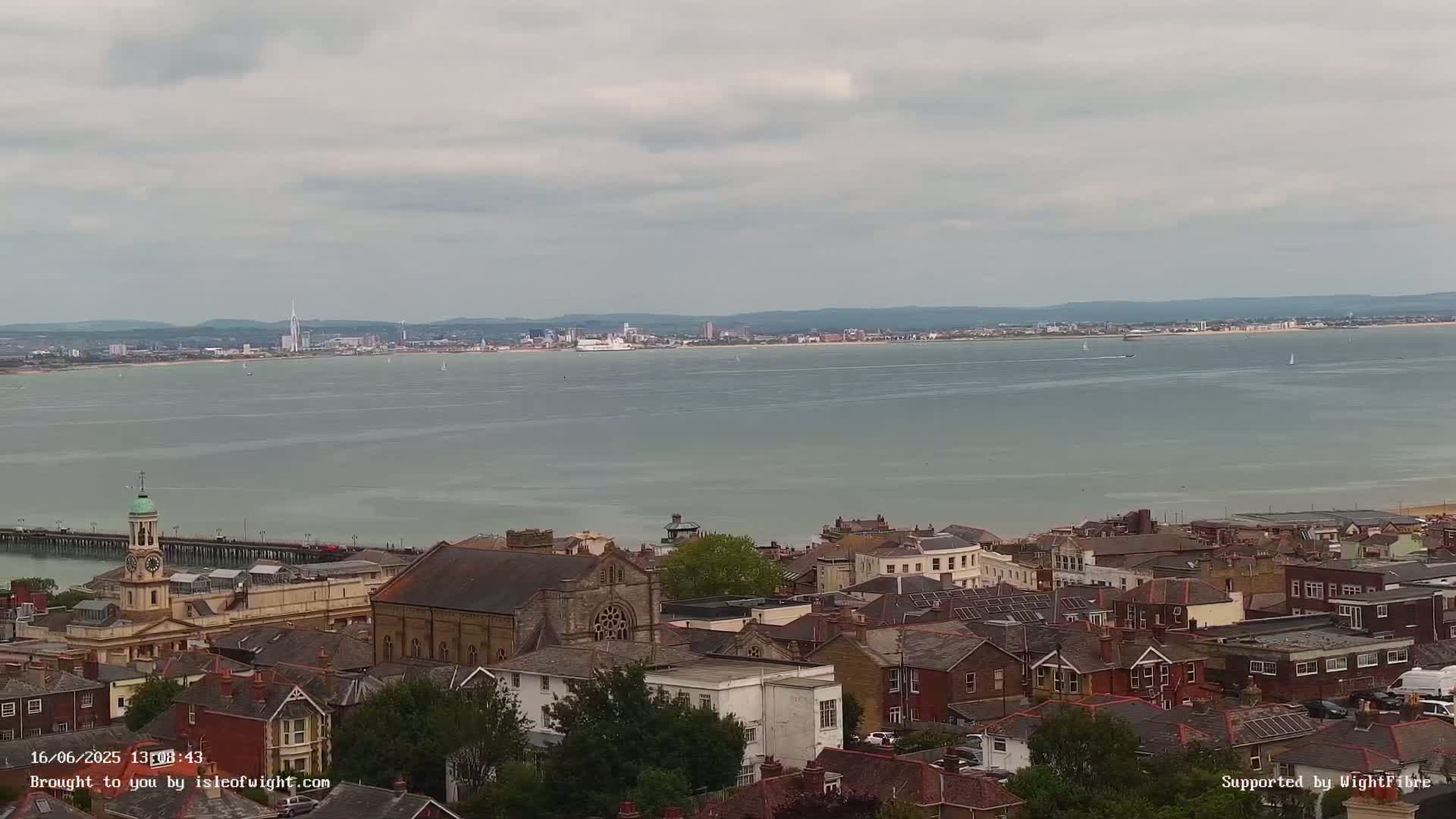 An overcast day reveals a coastal town with a pier, a church, and many houses, overlooking a calm body of water and a distant city skyline.