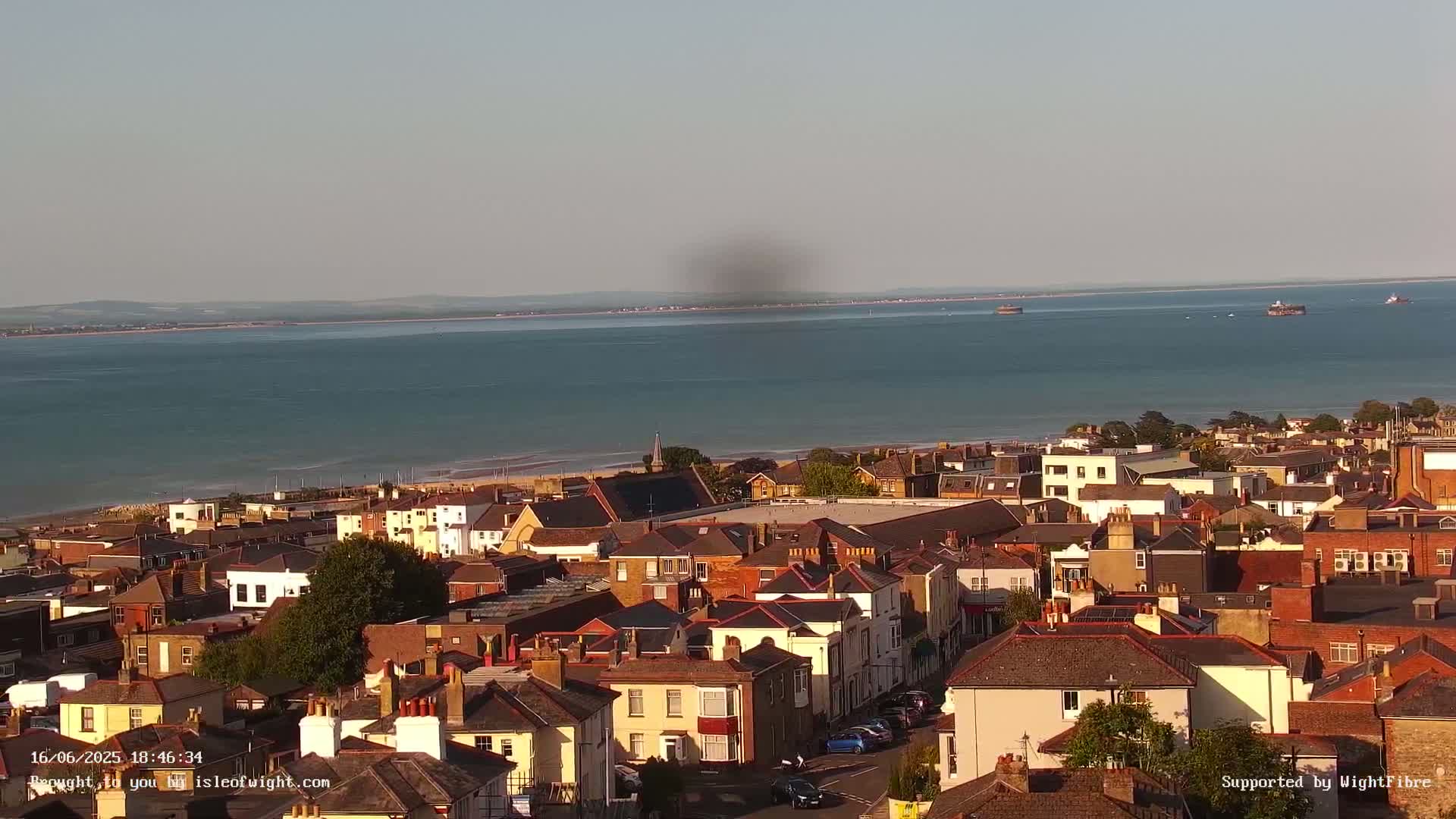 An aerial view shows a coastal town on a sunny day, with a calm body of water and a distant shoreline visible in the background.