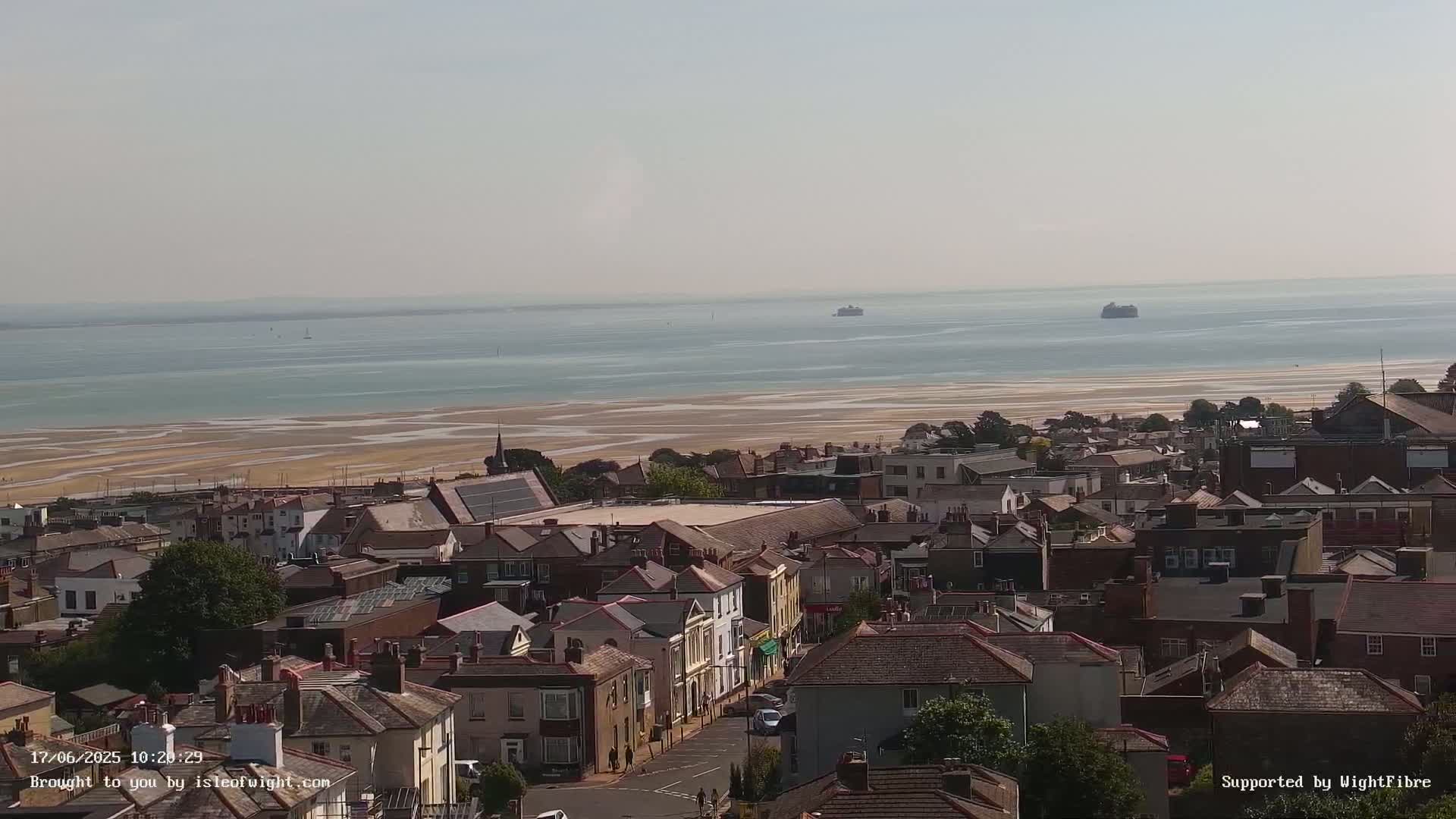 A coastal town with many brown-roofed houses is visible, overlooking a wide expanse of calm, light-blue water and a sandy beach under a hazy sky.