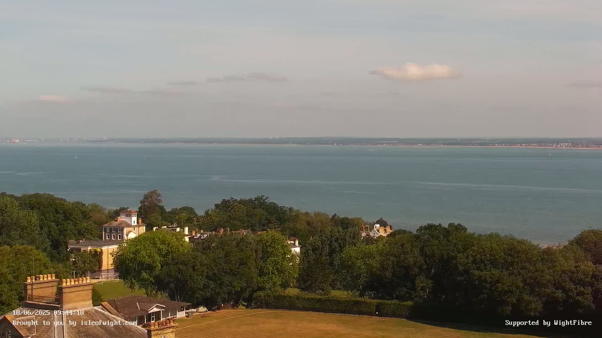 A tranquil, sunny day reveals a vast expanse of calm water stretching to a distant shoreline, overlooked by a verdant hillside dotted with houses and trees.