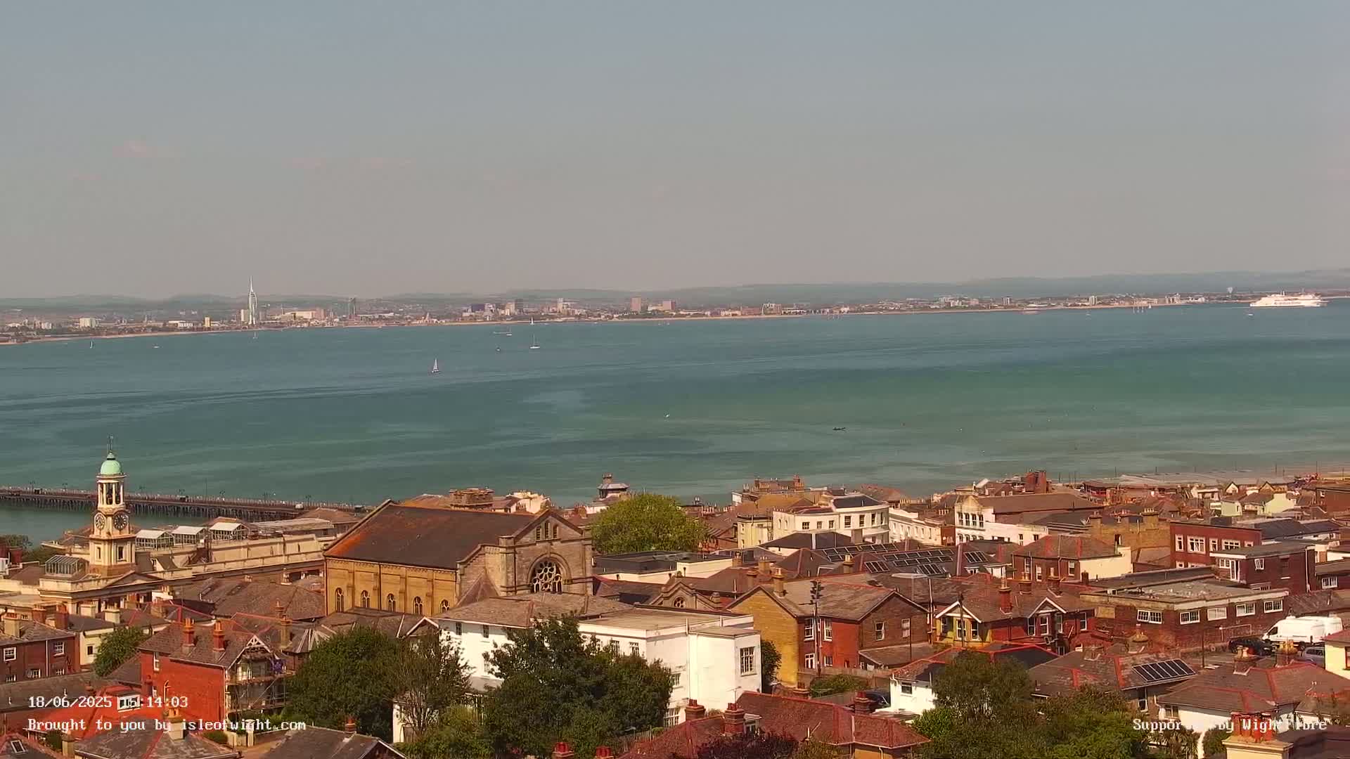 A coastal town with a mix of architectural styles overlooks a calm, teal-colored body of water on a sunny day, with a distant cityscape visible on the horizon.