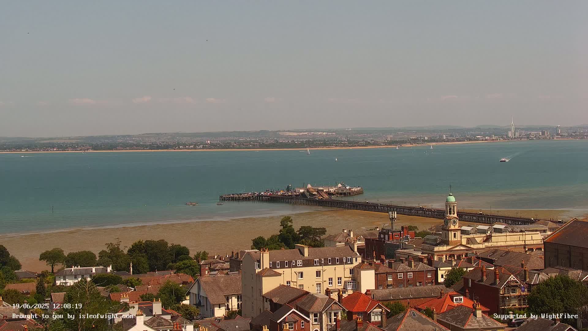 A seaside town with a long pier extending into a calm, teal-colored body of water under a mostly clear sky.