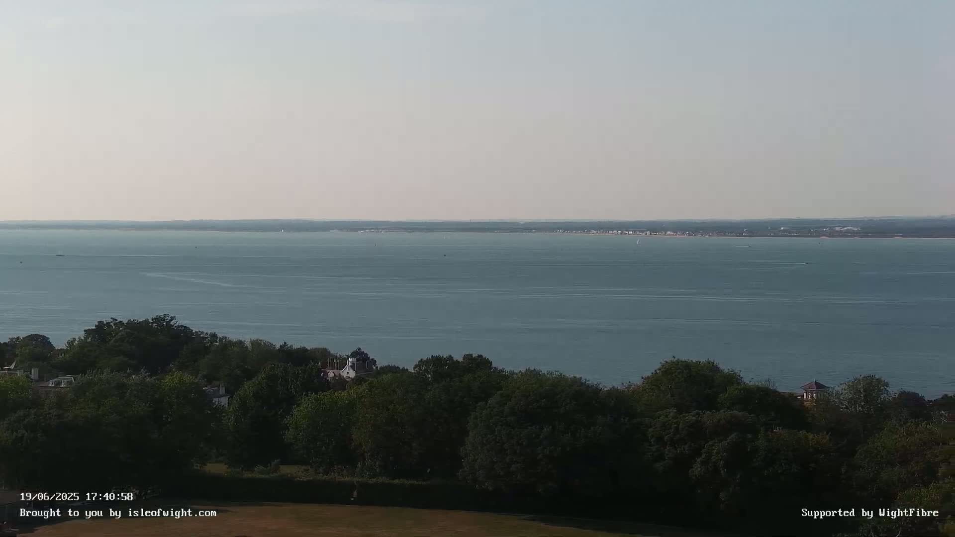 A calm, expansive body of water stretches to the horizon, under a clear sky, viewed from a high vantage point overlooking a line of dark green trees.