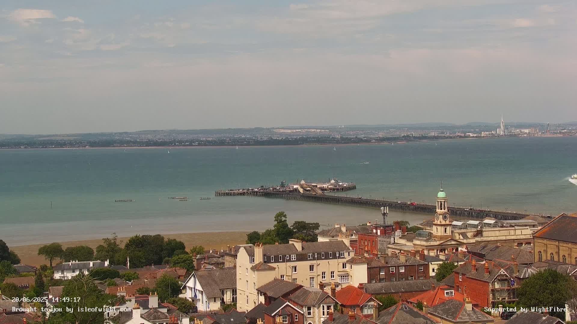 An elevated view shows a town overlooking a pier extending into a calm, teal sea under a partly cloudy sky.
