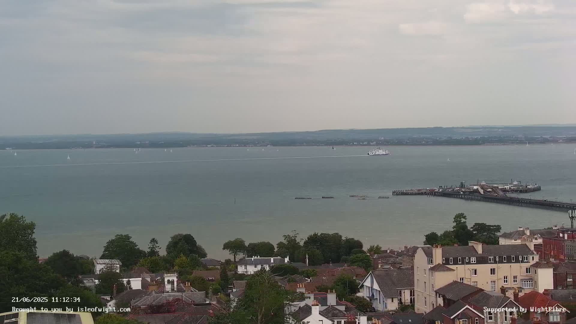 A ferry and sailboats are visible on a calm, overcast day across a body of water from a high vantage point overlooking a town with houses and a pier.