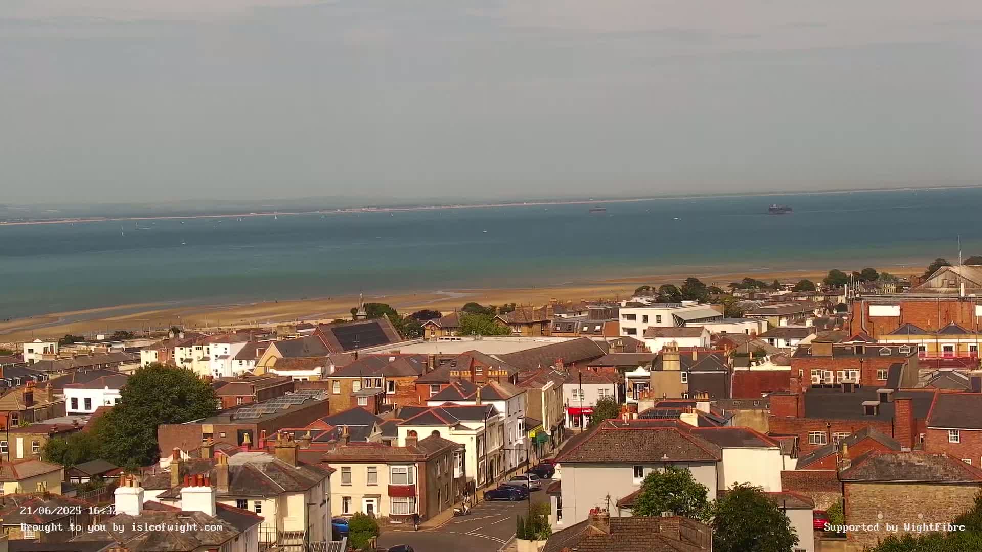 A coastal town with mostly brick buildings overlooks a calm, blue-green body of water and a wide sandy beach under a hazy sky.