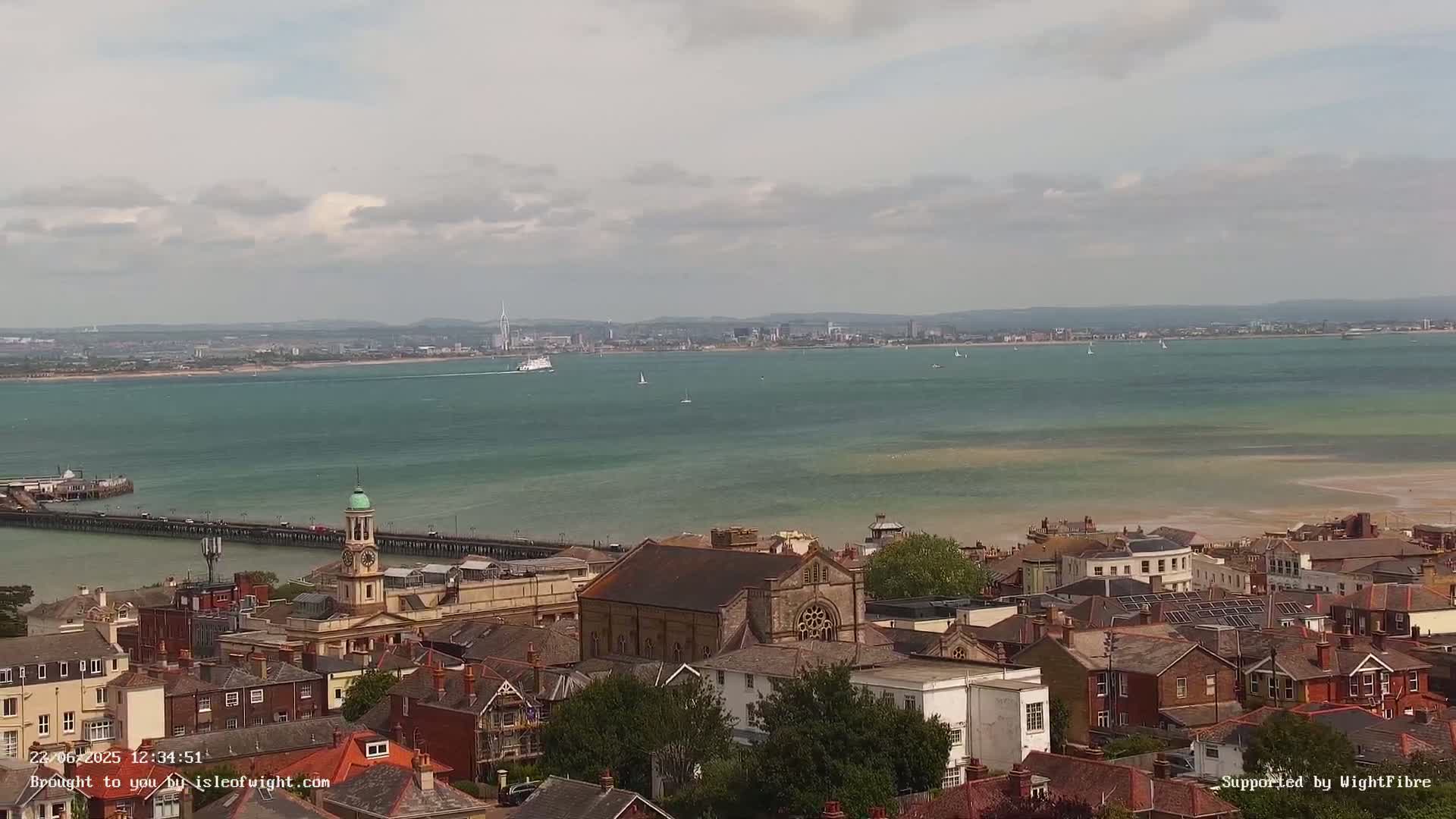 An elevated view shows a town with a clock tower and pier overlooking a calm body of water with a distant city skyline under a mostly cloudy sky.