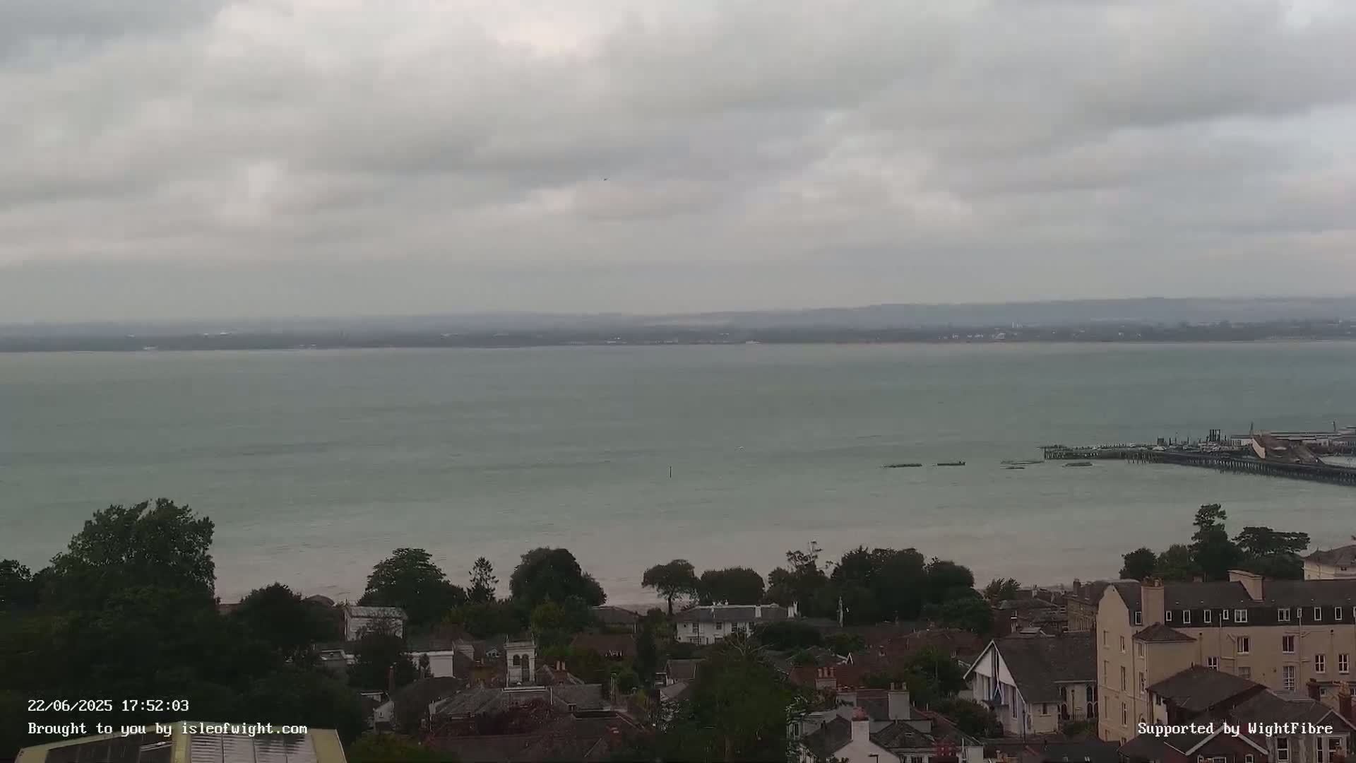 A wide shot of a calm body of water under an overcast sky, with a town and pier visible in the foreground.
