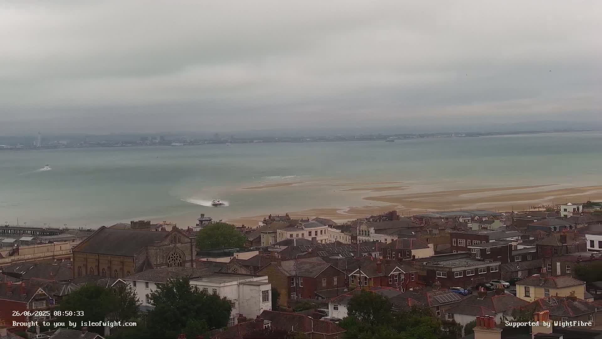 A hovercraft travels across a body of water toward a town under an overcast sky.