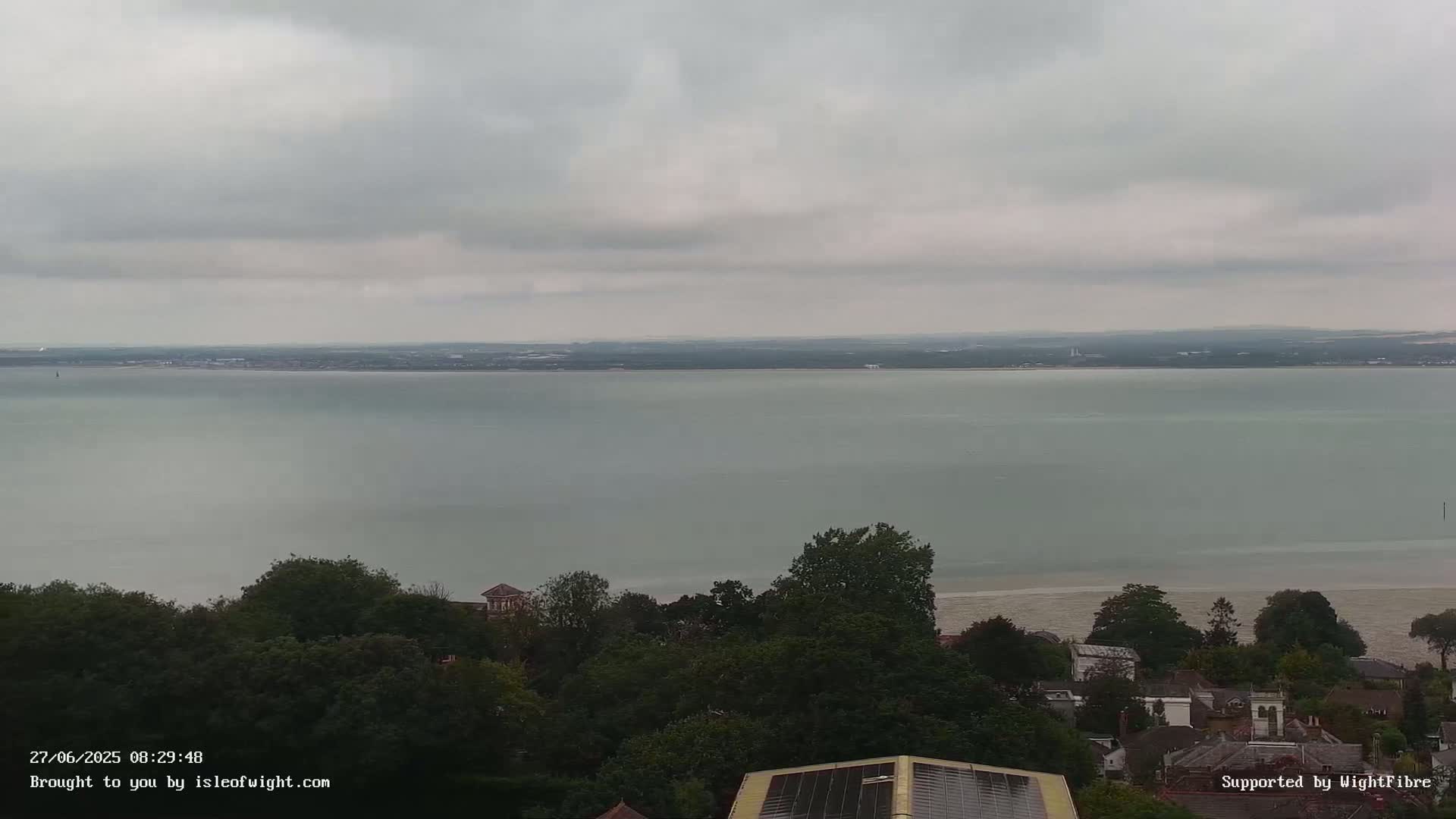 An overcast day shows a wide expanse of calm water stretching to a distant shoreline, overlooked by a line of trees and buildings.