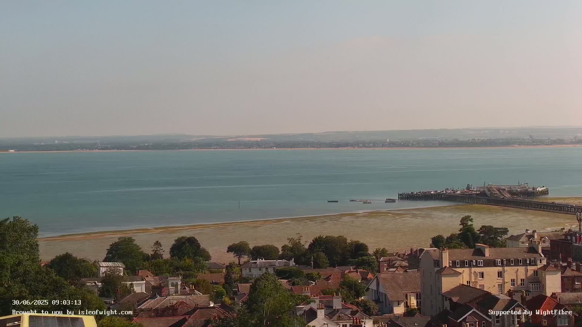 An aerial view shows a coastal town on a sunny day, overlooking a calm body of water with a pier extending into it.