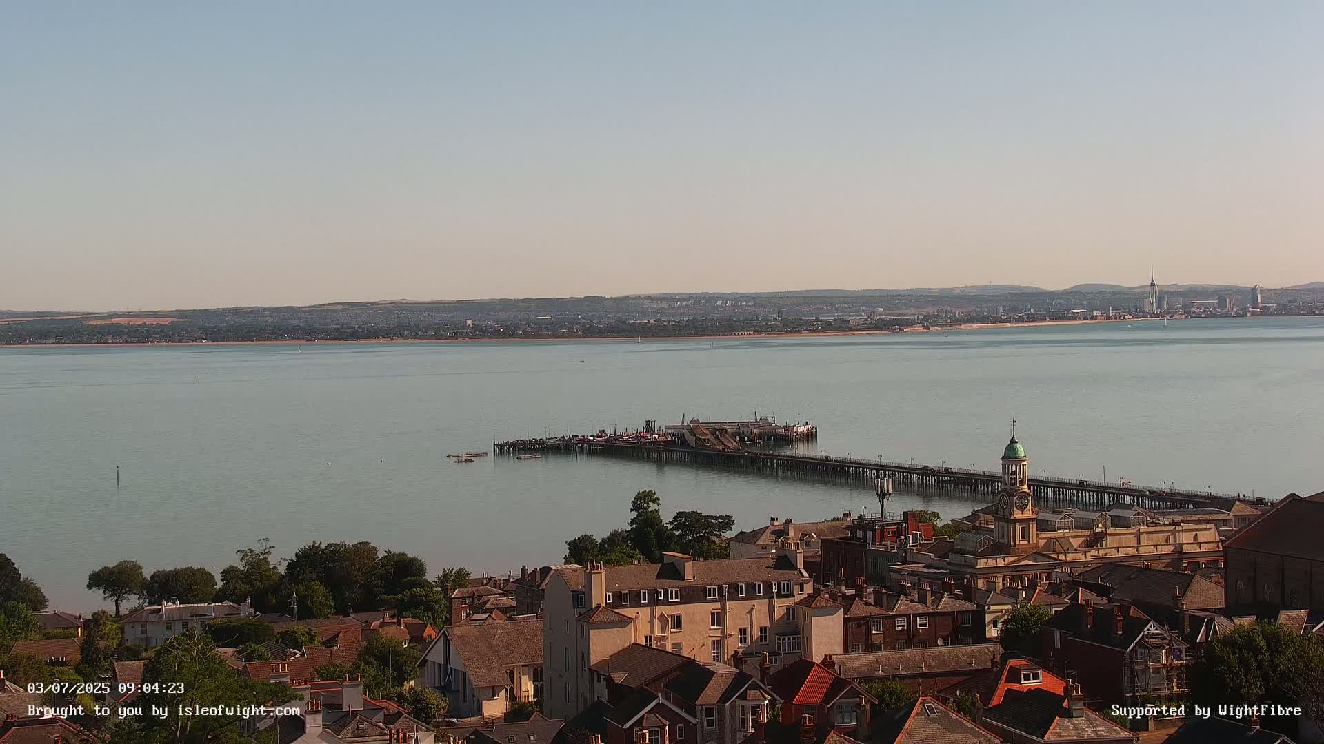 A long pier extends into a calm body of water, overlooking a town with buildings of varying heights and styles under a clear, bright sky.