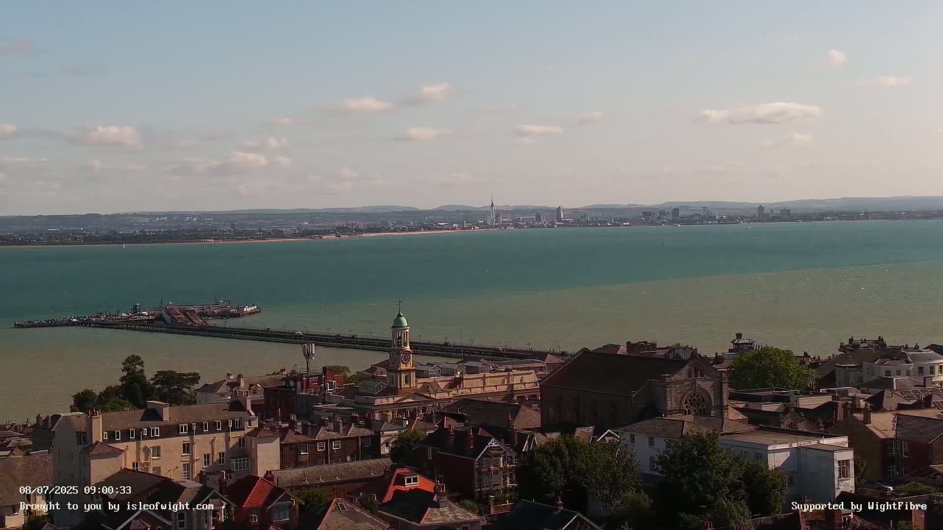 A coastal town with a pier and clock tower is visible, overlooking a calm body of water with a distant city skyline under a partly cloudy sky.