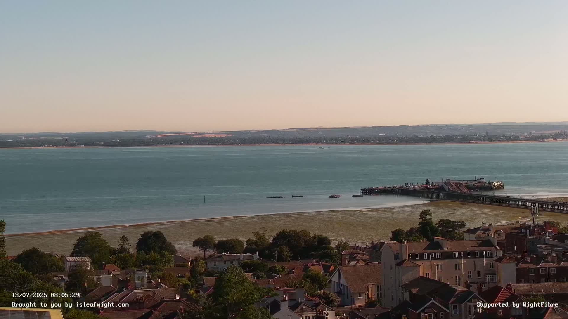 A long pier extends into a calm, teal sea under a clear, light-pinkish sky, overlooking a town of brown-roofed houses.