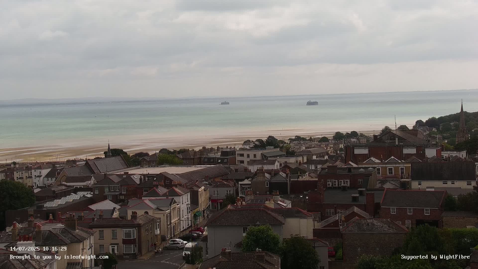 A coastal town with many houses and a calm, light-green sea under an overcast sky is seen from an elevated viewpoint.