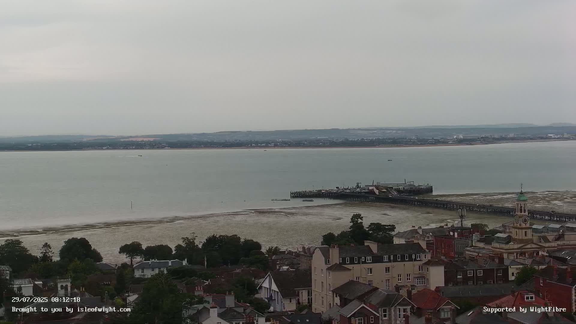 A coastal town with a long pier extends into a calm, overcast body of water, with a distant shoreline visible on the horizon.