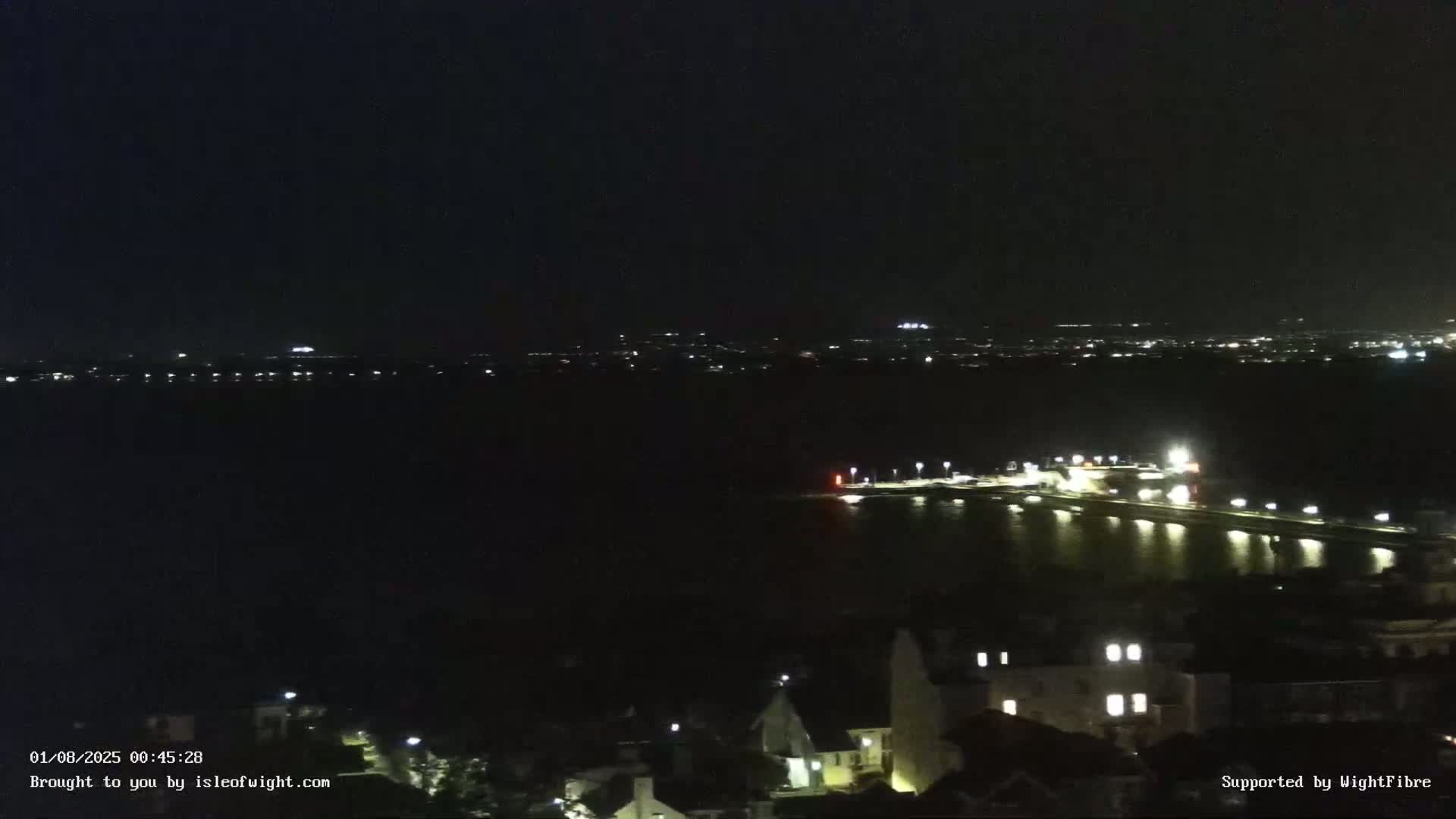 A nighttime view shows a dark body of water with a lit waterfront and distant city lights under a clear, dark sky.