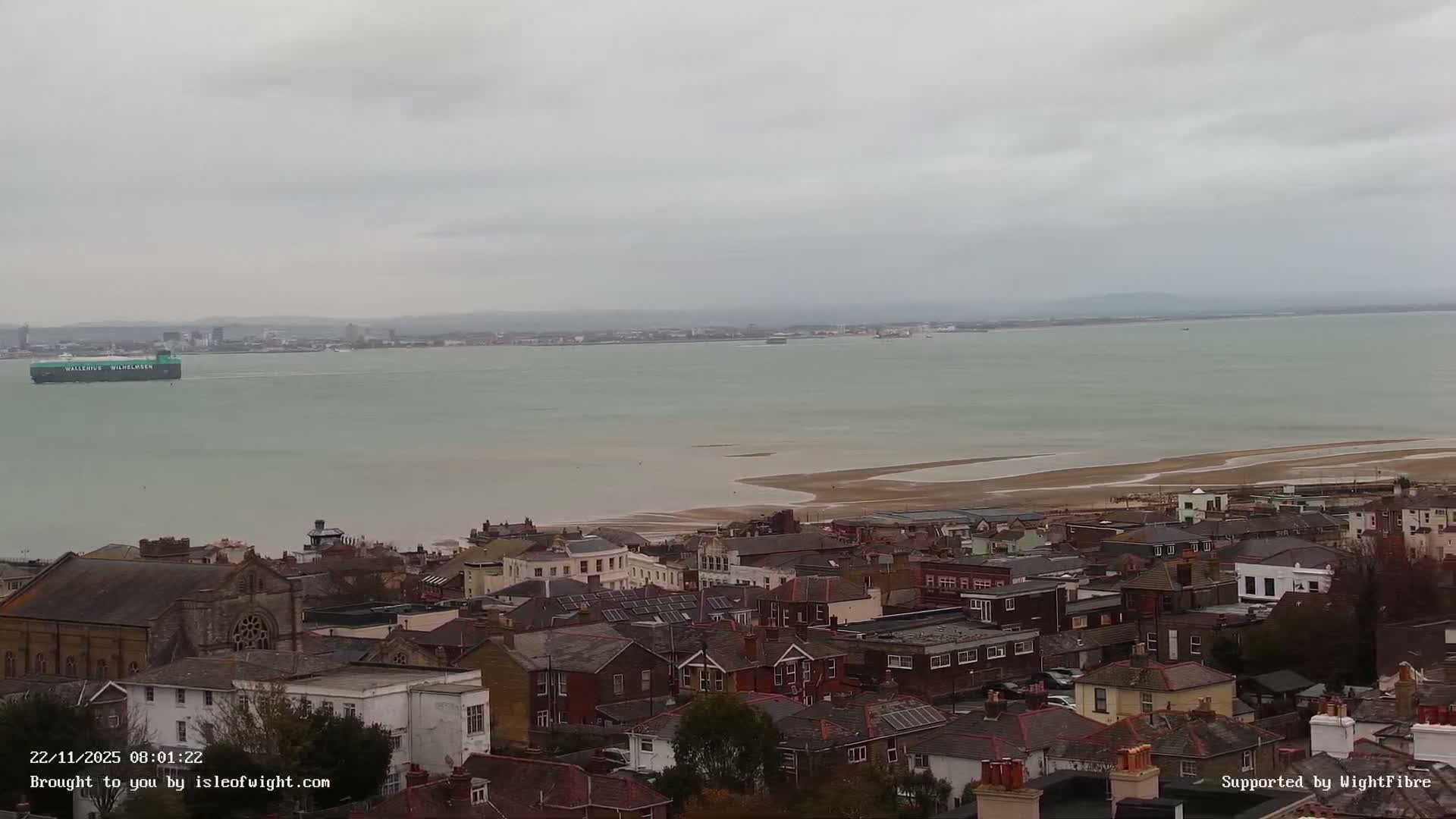 An elevated view on an overcast day shows a dense coastal town with varied rooftops, a sandy beach, and a large green cargo ship crossing wide, calm water towards a hazy distant city.