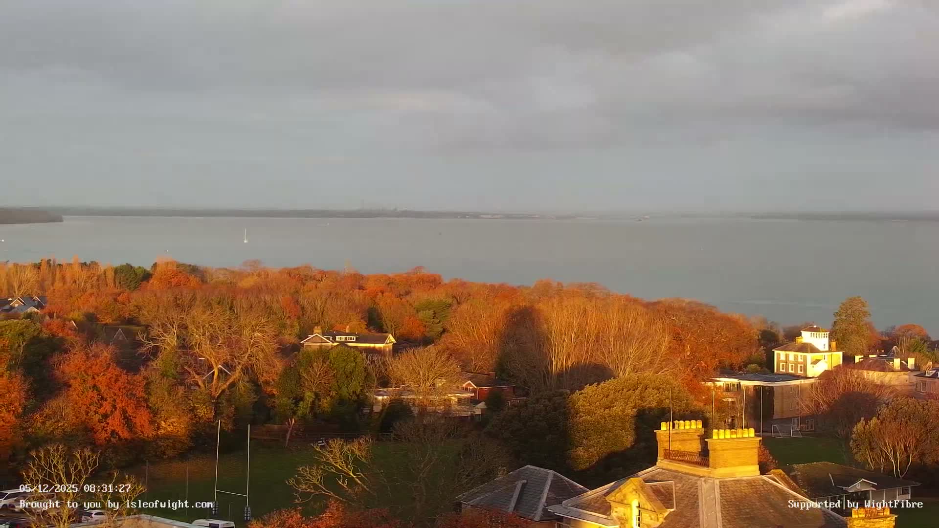On a partly cloudy autumn day, an elevated vista showcases a wide bay bordered by hillsides bursting with vibrant orange and yellow foliage and interspersed residential buildings, all under a grey sky with sunlight highlighting sections of the landscape.