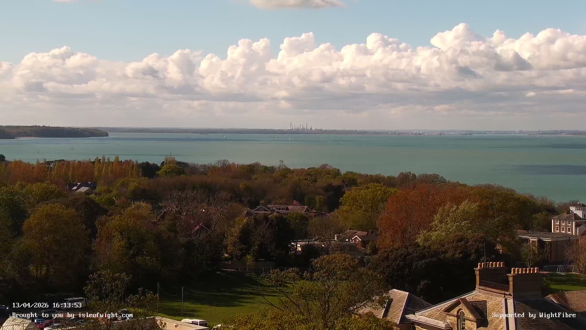 On a partly cloudy autumn day, an elevated vista showcases a wide bay bordered by hillsides bursting with vibrant orange and yellow foliage and interspersed residential buildings, all under a grey sky with sunlight highlighting sections of the landscape.