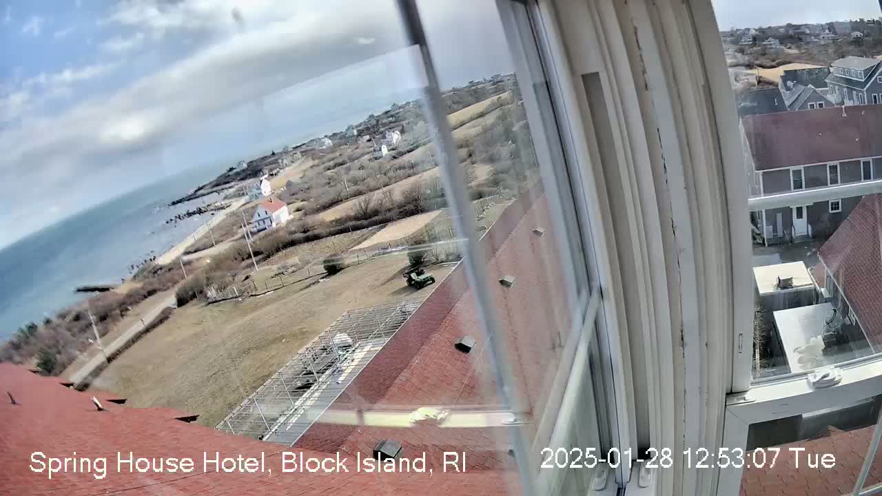 A view from a window shows a partly cloudy day overlooking a coastal town with a body of water and houses, including a red-roofed building partially visible in the foreground.