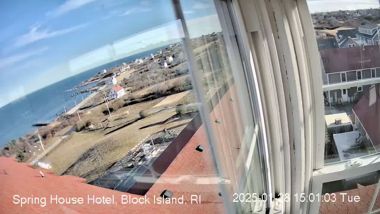 A high-angle view from a window shows a coastal town on a sunny day, with a body of water visible in the background.