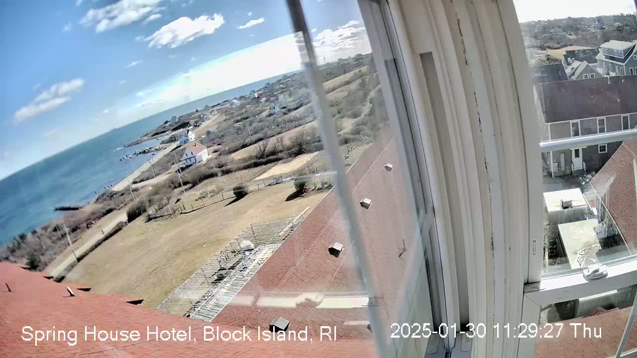 A view from a window shows a mostly sunny day overlooking a coastal town, ocean, and a red roofed building.