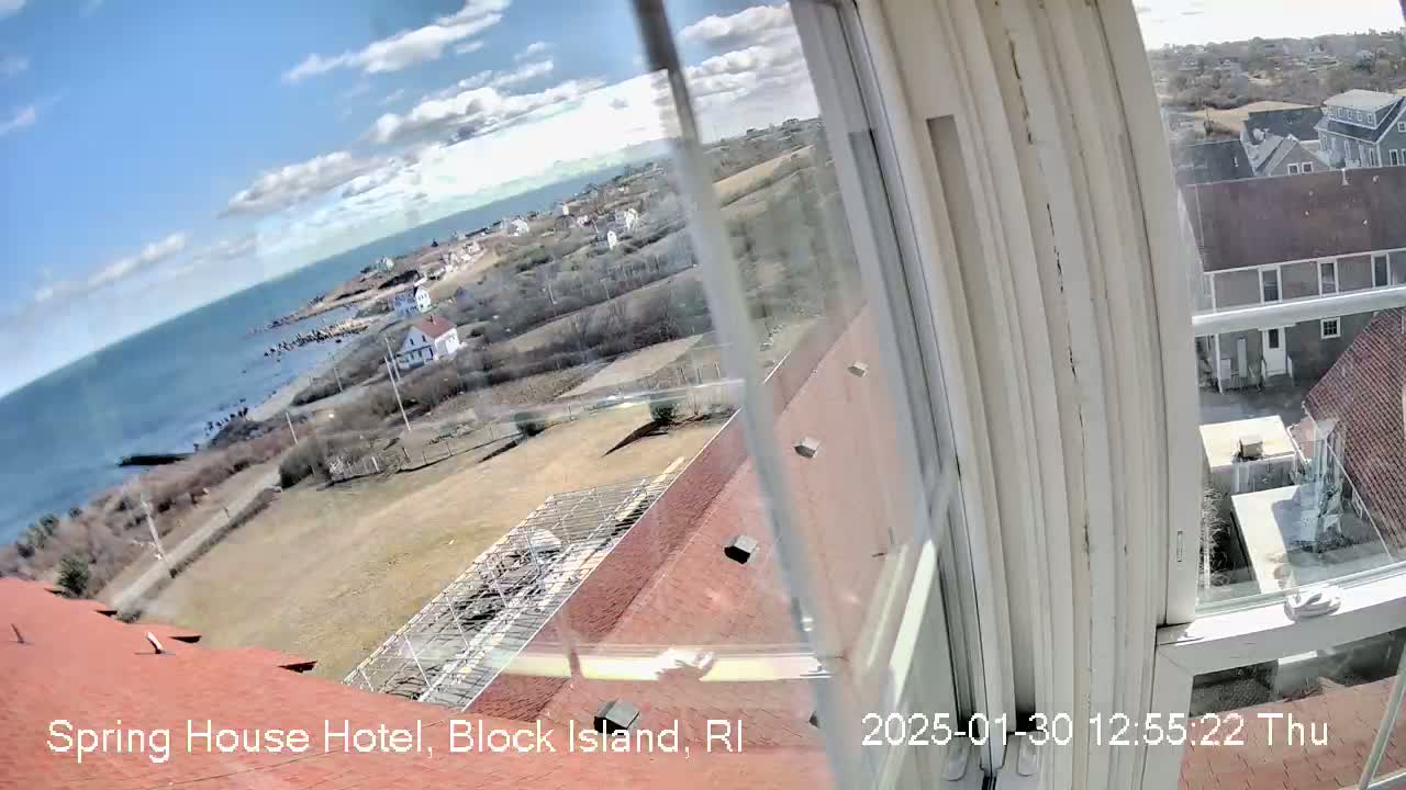A view from a window shows a partly sunny day overlooking a coastline with buildings, a grassy area, and the ocean.