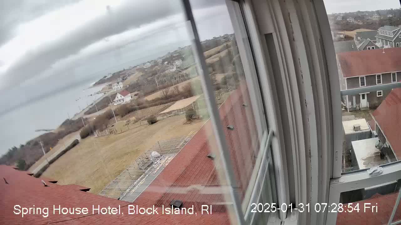 A view from a window shows a coastal town on an overcast day, with red-roofed buildings and a body of water visible in the distance.